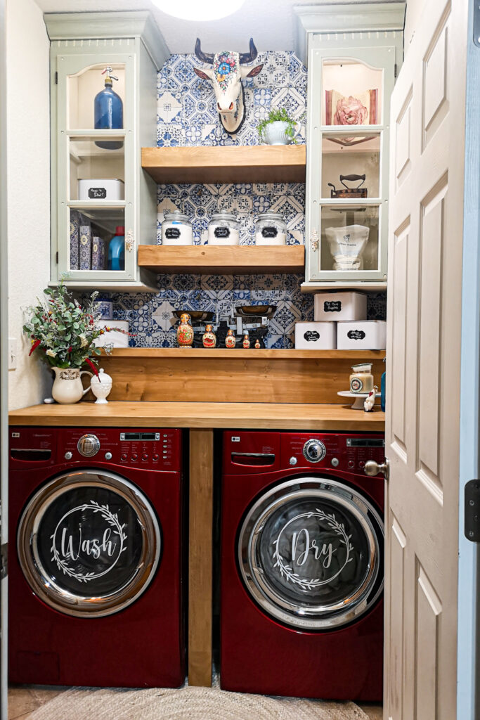 Stunning Laundry room with red front loading washer and dryer. A beautiful wood folding counter made from an old dining table. Blue and white French tiled wall paper is a back drop to two stunning grey green glass door cabinets that used to be an old china hutch. Floating shelves reach between the two cabinets with a surprise in the lower one as a hidden drawer pulls out with a drying rack in it.